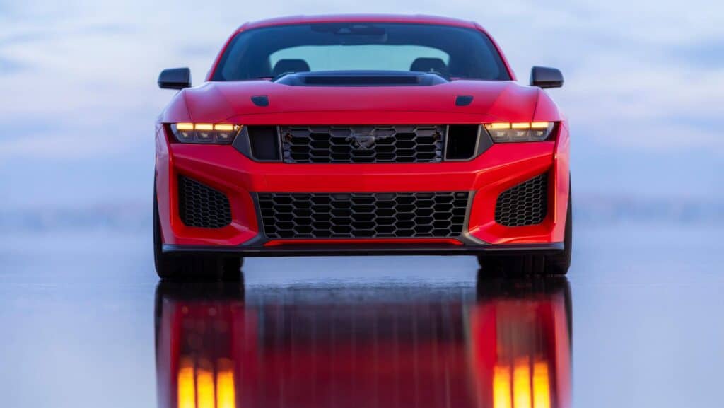 Red sports car front view with sleek design and illuminated headlights, reflected on a wet surface.