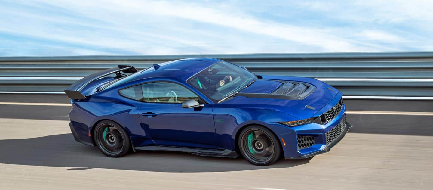 Blue sports car driving on a road with a metal guardrail. Clear sky in the background.